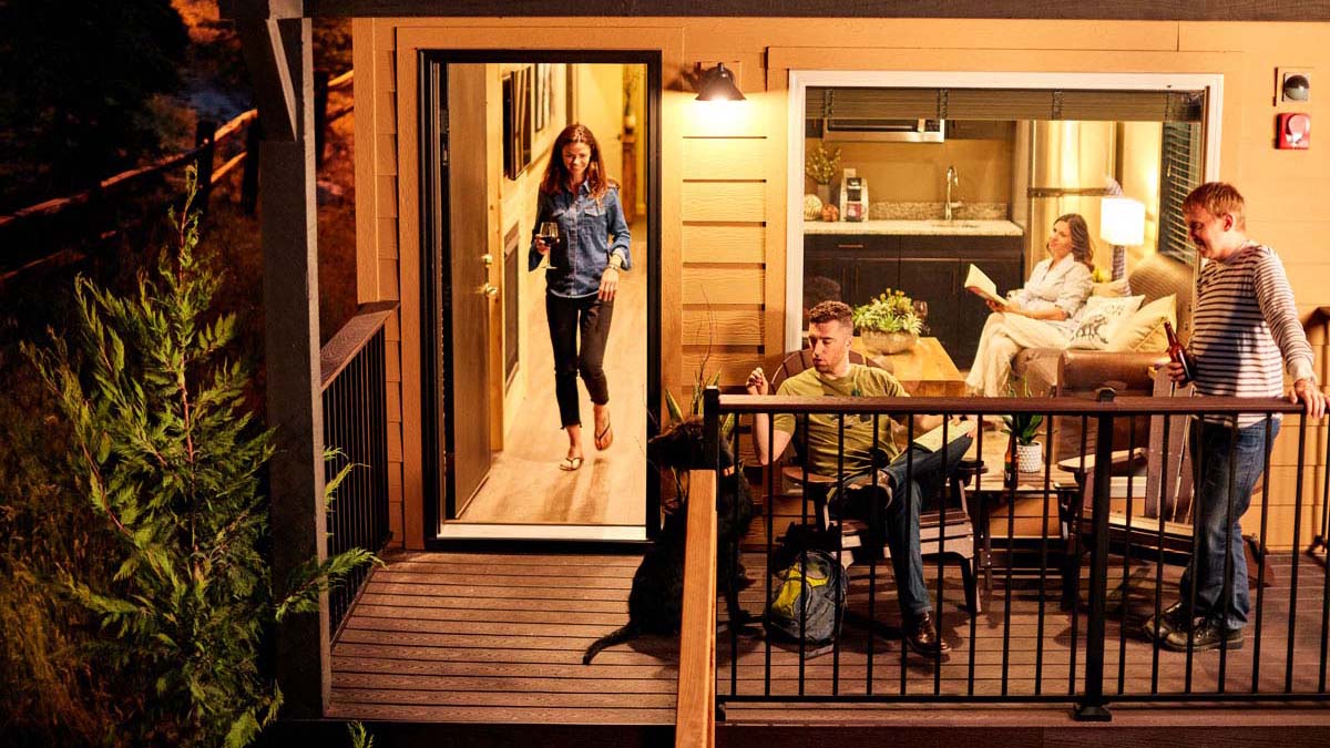 In central Mariposa County, a small group finds comfort in the inviting space of a tiny house designed with dog enthusiasts in mind. The scene captures someone gently holding their dog on the wooden deck, while another person steps through the nearby door. Inside, two more people are seated comfortably, absorbed in conversation or leisure. This quaint retreat offers pet-friendly accommodations and an appealing interior that serves as an ideal base for exploring Yosemite. Outside, a single tree provides natural shade alongside the deck.