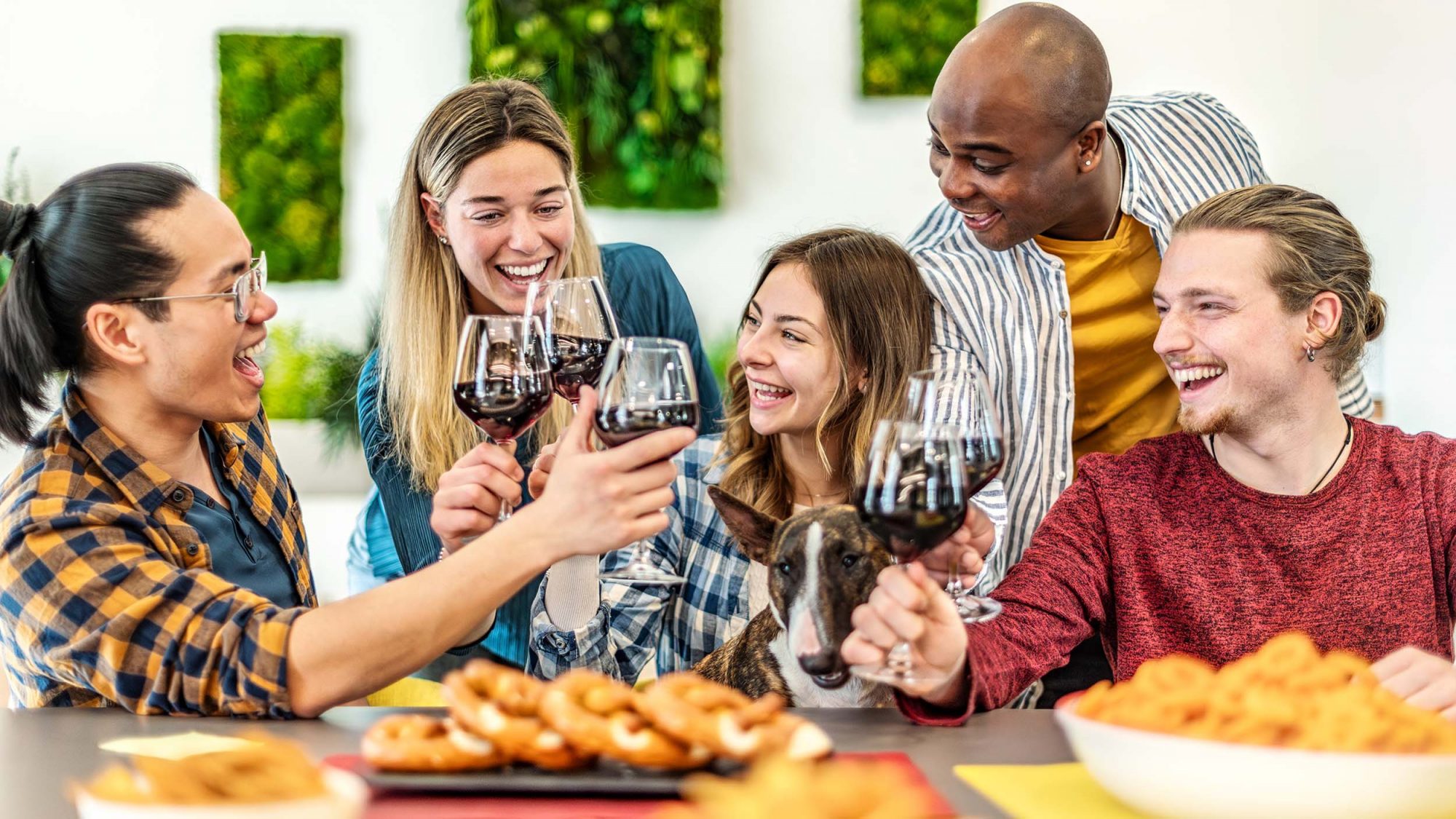 Five friends gather around a table, each holding a glass of red wine. They raise their glasses in a toast, enjoying the generous discounts on their preferred bottles. A dog, possibly representing Pawsport for Napa Humane, lies contentedly at their feet. The table is spread with an assortment of snacks and the wall behind them is decorated with eye-catching green artwork.