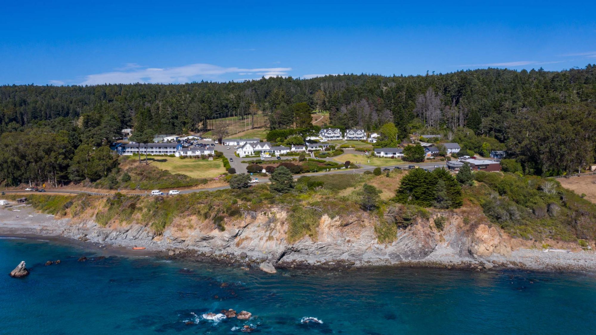 The aerial view captures the stunning coastal landscape near Little River Inn, where a rugged shoreline meets the expansive blue ocean. A collection of white buildings is tucked amidst a canopy of green trees and open grassy spaces on a gently sloping hillside. The vibrant, cloudless sky provides an ideal backdrop for the upcoming Pup Palooza 2025 events, promising an inviting setting for both dogs and their owners.