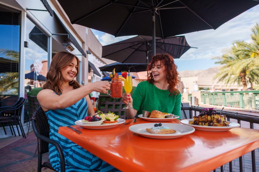 In the sunlit atmosphere of Greater Palm Springs, two women sit at a table outdoors, their glasses meeting with a subtle clink. Around them, large black umbrellas provide shade, making it an ideal spot for dining with their dogs. On their plates are servings of both pancakes and salad. As they eat, they take in the sight of palm trees swaying gently against a bright blue sky—a picturesque backdrop that complements their carefully planned day with their pets.