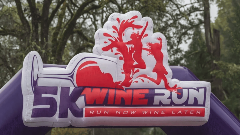 An inflatable archway stands prominently, featuring the "5K Wine Run" logo, a design that includes sleek images of runners alongside a glass of wine. Below the logo, it reads, "Run Now Wine Later," enticing participants with its promise. Set amidst the picturesque backdrop of Nelson Family Vineyards, this event offers a unique opportunity to enjoy an invigorating run or leisurely walk surrounded by nature and followed by wine tasting.