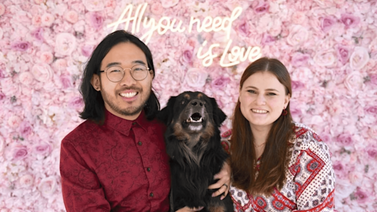 A couple stands in front of a vibrant wall adorned with pink blooms, their expressions warm and content. Between them sits a joyful black and brown dog, its eyes bright with excitement. Above this lively scene, a neon sign declares, "All you need is furever love." The trio is dressed in vividly patterned clothing that highlights the spirited nature of animal rescue and pet care.