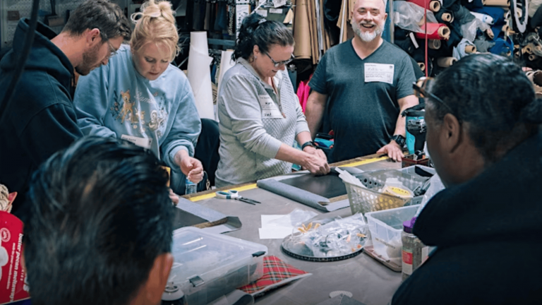 A group gathers around a table cluttered with colorful craft supplies, each person intently focused on their project. Shelves brimming with sustainable materials, such as ropes and fabrics in various textures, line the background. One individual stands slightly apart, demonstrating techniques to the others eagerly crafting personalized toys for their pets at this hands-on Pet Toy Workshop.