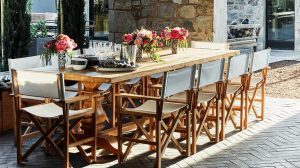 A spacious wooden dining table is arranged for guests, featuring neatly placed plates and glasses alongside floral centerpieces from Stewart Cellars. Director-style chairs with white canvas seats and backs offer comfortable seating around the table. This welcoming setting is located on a patio surrounded by sturdy stone walls and tall trees, providing a peaceful atmosphere for a meal.