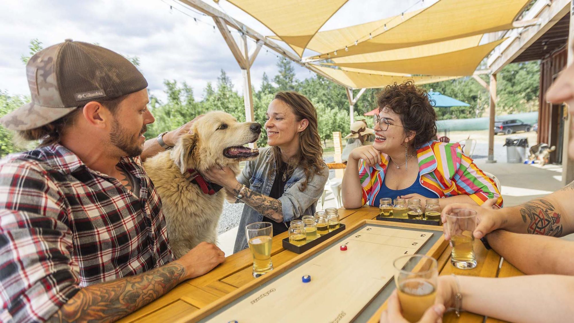 Four people sit around an outdoor table, engaged in a tabletop game with drinks beside them. Laughter and smiles fill the air as they enjoy each other’s company. A large dog lies contentedly nearby, savoring his meal of Bone Appétit gourmet pet food. The woman in the middle leans down occasionally to give him affectionate pats. The scene is lit by natural sunlight, softened by overhead shade canopies that provide a comfortable atmosphere for both humans and their furry companion.