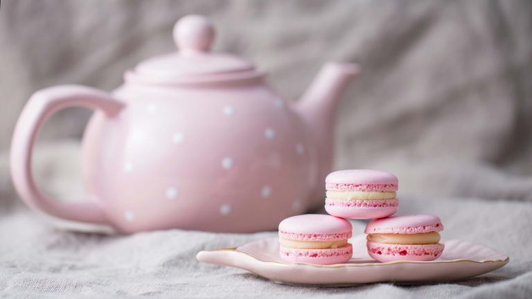 A pink teapot with white polka dots rests on a soft beige cloth, setting the scene for an afternoon tea. Three pink macarons are neatly stacked on a wavy-edged ceramic dish in front of it, forming a simple yet refined presentation.
