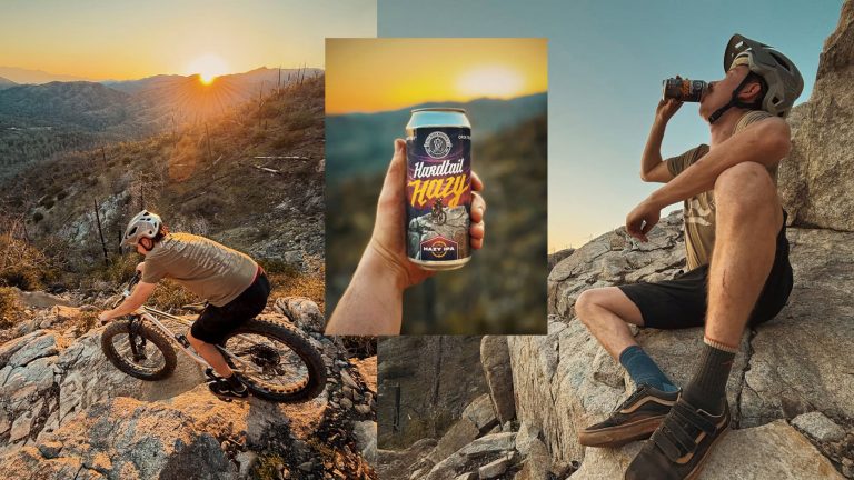 A cyclist wearing a helmet and protective gear navigates rugged trails on a mountain bike as the sun sets, casting dramatic hues across the rocky landscape. They come to a stop and take a refreshing sip from a can prominently featuring the label of "Hardtail Hazy IPA." The scene captures the essence of an adventurous ride, perfectly timed with the celebration of the IPA Release weekend, set against an awe-inspiring backdrop that invites exploration.