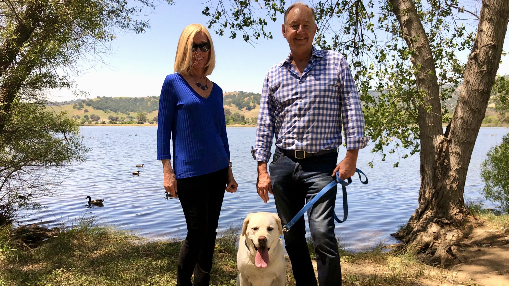 A couple spends the day by the lake with their dog securely on a leash. The woman wears a blue top, and her companion opts for a checkered shirt. Ducks glide across the water at an unhurried pace. Trees flanking the scene suggest a tranquil setting under sunny skies, perfect for enjoying time outside with their pet.