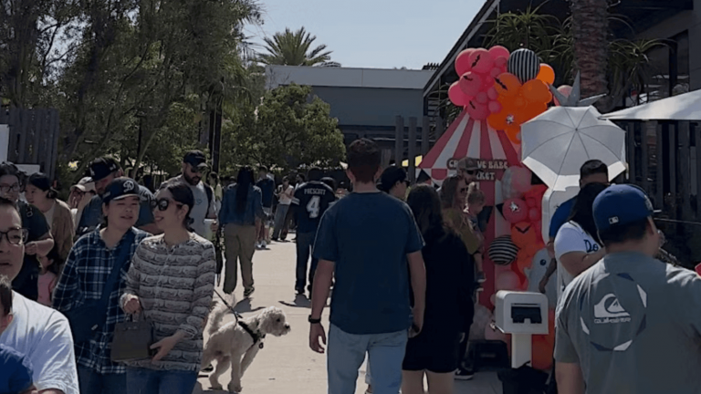 At the heart of a lively pop-up market, people stroll and interact, their voices mingling with the rustle of leaves from nearby trees. A dog trots alongside its owner, tail wagging with excitement. They pause in front of a vibrant booth—a splash of color beneath a striped canopy—where balloons sway gently in the breeze. Behind this scene rises an elegantly structured building, framing the bustling activity below against its stately backdrop.