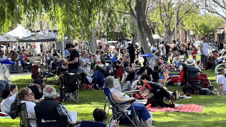 A diverse crowd settles on lawn chairs and blankets scattered across the grassy expanse of a park during Bay Area's SpringFest. Towering trees offer shade, casting dappled patterns on the ground. In the background, white tents stand in neat rows, housing various attractions and vendors. The sun shines brightly overhead, adding warmth to this vibrant community gathering where attendees savor the lively atmosphere with their dogs by their side.
