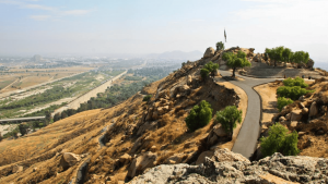 A path weaves its way to the summit of Mount Rubidoux, a stony hill dotted with patches of green shrubs and crowned by a tall flagpole. This well-known spot in Riverside offers sweeping views: an extensive vista of roads crisscrossing through fields, all set against the backdrop of California's distant mountain ranges beneath a sky partially covered with clouds.