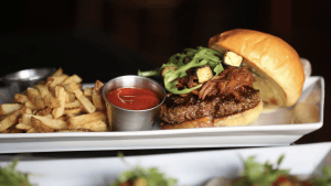 A gourmet burger rests on a rectangular white plate, its lustrous golden bun crowned with crisp greens and crunchy croutons. Beside it lies a serving of freshly cooked fries, their warm aroma inviting. A small metal cup of vibrant red ketchup completes the ensemble, ready for dipping.