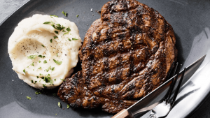 At Paul Martin's American Grill, you can savor a ribeye steak grilled to perfection, showcasing distinct grill marks. It is accompanied by creamy mashed potatoes and garnished with chopped herbs for added flavor. The meal is presented on a sleek dark plate, with a knife and fork placed neatly beside it for your convenience.