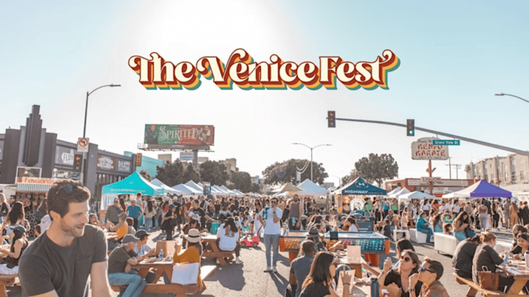 The outdoor festival hums with activity as people move through the vibrant scene, savoring food and engaging in various activities. Tents and booths stretch across the area, each offering something unique and enticing. A prominent sign overhead reads "Venice Love Fest 2025," while the clear sky and bright sunlight bathe everything in a cheerful glow.