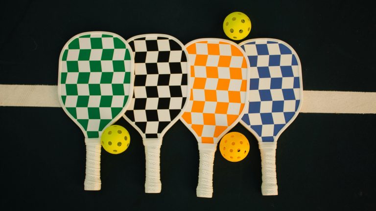 Four pickleball paddles lie side by side, each displaying a checkered design in distinct colors: one green, another black, the third orange, and the last blue. Next to each paddle rests a bright yellow pickleball. They are arranged against a dark backdrop marked by a singular horizontal white line. This setup is organized for the Pickle Paws benefit tournament, an event welcoming dog lovers and their canine companions.