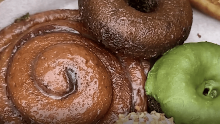 At the San Diego Night Market, you can explore a dessert platter that offers a variety of vegan treats, perfect for anyone with a sweet tooth. On this visually appealing white plate, you'll find a chocolate-frosted doughnut sitting next to one with a distinctive green glaze. A cinnamon roll promises warm spices and sweetness in every bite, while clusters of oats add texture and depth. A crunchy pretzel stick provides a salty contrast to the other sweet offerings. This assortment might tempt humans more than dogs—but nearby food stalls often have pet-friendly options so furry companions can have their own treat too!.