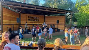 A live band plays on a wooden stage marked “Trouble Makers” at Troublemakers Beer Garden. People with dogs gather around the stage, some standing close to the railing with glasses of craft beer in hand. Several audience members dance or sway along to the music, while others watch from picnic tables or benches nearby. String lights hang from tree branches over the gathering, casting soft light across the garden and footpaths where dogs rest at their owners’ sides or sniff along the grass.