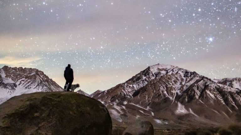 A person stands on a broad granite boulder in Buttermilk Country, watching the stars over the snow-covered Sierra Nevada peaks. Scattered shrubs and short grasses dot the ground nearby.