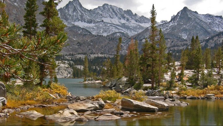 Lake Sabrina is a clear alpine lake bordered by boulders and tall pine trees. The water’s edge is rocky, with patches of low bushes and tufts of grass. Beyond the shore, steep mountain peaks rise, dusted with late snow and framed beneath an overcast sky. The trails around the lake offer opportunities for leashed walks along the shoreline and up into the surrounding mountains.