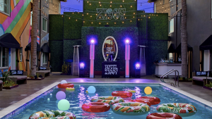 A rectangular outdoor pool filled with inflatable floats and bright plastic balls sits in the center of Hotel Ziggy’s courtyard. The pool faces a stage marked "Hotel Ziggy," where large speakers stand on both sides. Overhead, string lights stretch across the space, casting a glow over the modern hotel surroundings.