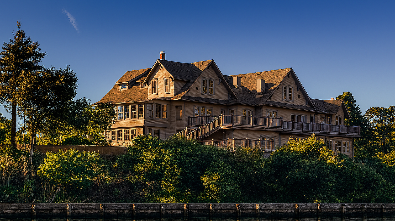 A vibrant dog park bathed in sunlight features abundant greenery with mature trees, offering ample shade for leisurely outdoor activities. In the distance is a well-equipped playground that presents an additional activity area. An inviting picnic table spreads out nearby, ideal for enjoying meals al fresco or taking relaxing breaks from play. A neatly-placed trash bin aids in maintaining the park's cleanliness for everyone's enjoyment. Dominating the scene is a generously wide dirt path gracefully curving through the expanse of grass, promising exploratory walks and lively fetch games.