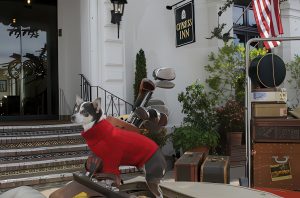 A small dog in a red sweater sits on the hood of a vintage car parked outside the Cypress Inn, a hotel in California that welcomes pets. Several suitcases and golf bags rest beside the car. An American flag hangs overhead.