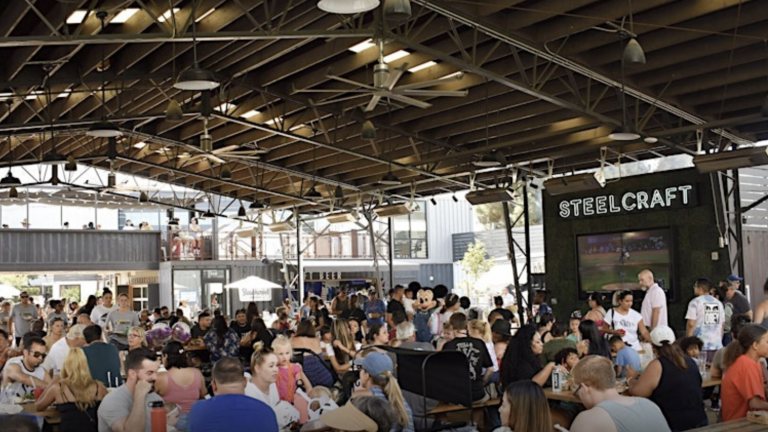A vibrant dog park bathed in sunlight features abundant greenery with mature trees, offering ample shade for leisurely outdoor activities. In the distance is a well-equipped playground that presents an additional activity area. An inviting picnic table spreads out nearby, ideal for enjoying meals al fresco or taking relaxing breaks from play. A neatly-placed trash bin aids in maintaining the park's cleanliness for everyone's enjoyment. Dominating the scene is a generously wide dirt path gracefully curving through the expanse of grass, promising exploratory walks and lively fetch games.