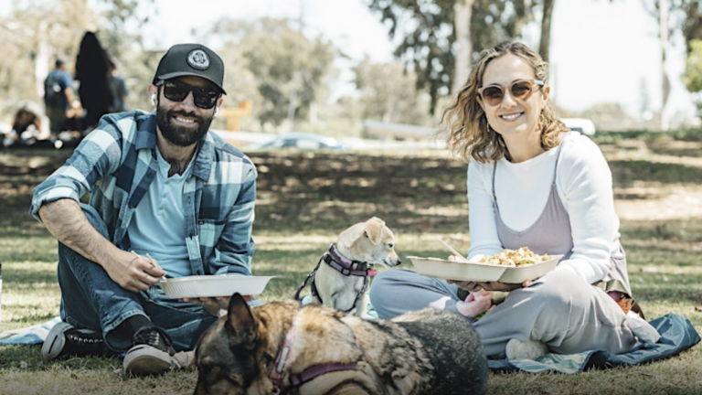 At a San Diego food festival, two people eat with their dogs, one lying and one sitting, on a blanket in a park.