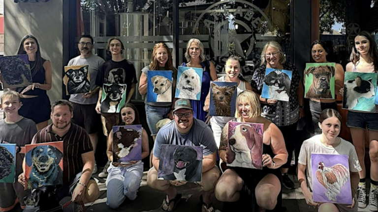 People at a Paint Your Pet event hold up their dog portraits outside, arranged in two rows near a building with large glass windows.