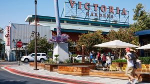 A vibrant dog park bathed in sunlight features abundant greenery with mature trees, offering ample shade for leisurely outdoor activities. In the distance is a well-equipped playground that presents an additional activity area. An inviting picnic table spreads out nearby, ideal for enjoying meals al fresco or taking relaxing breaks from play. A neatly-placed trash bin aids in maintaining the park's cleanliness for everyone's enjoyment. Dominating the scene is a generously wide dirt path gracefully curving through the expanse of grass, promising exploratory walks and lively fetch games.