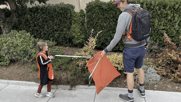 An adult and child in orange vests pick up trash with grabbers beside a sidewalk; green bushes line the area behind them.