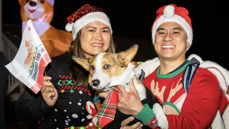 Festive holiday photo of a smiling couple with their dog, both wearing Christmas sweaters and Santa hats, celebrating Christmas at a winter event, with a large inflatable dog in the background.