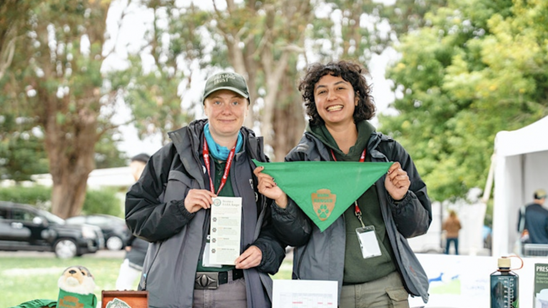 1. Smokey Bear banner with two smiling women outdoor at a wildlife conservation event, holding a green bandana with Smokey Bear logo, surrounded by trees and informational booths.