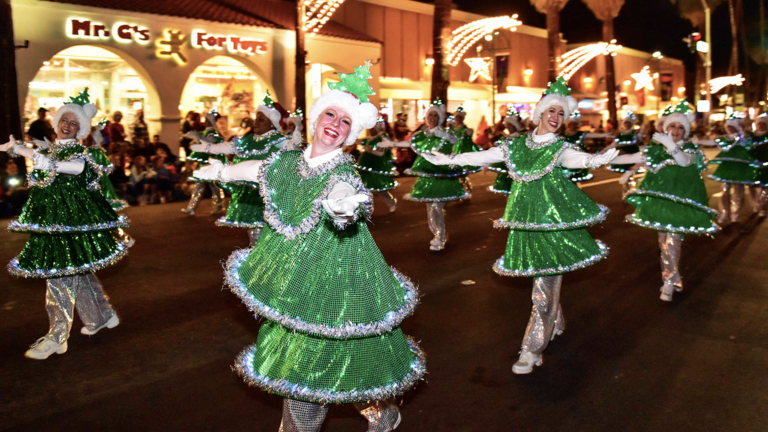 Colorful Christmas parade with women dressed in sparkly green holiday costumes, dancing and celebrating on a lively street illuminated by festive lights and holiday decorations.